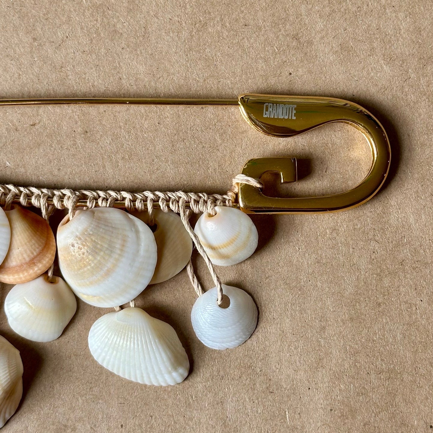 A gold safety pin brooch decorated with small white and beige seashells on a textured kraft paper background.