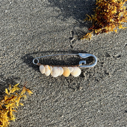 A silver safety pin repurposed as a beaded necklace with small white and beige shells, lying on dark gray sand with yellow seaweed in the corners.