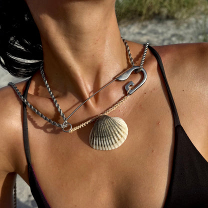 A close-up lifestyle shot of a woman at the beach wearing layered necklaces, including a silver safety-pin style necklace with a seashell pendant and a silver chain necklace.