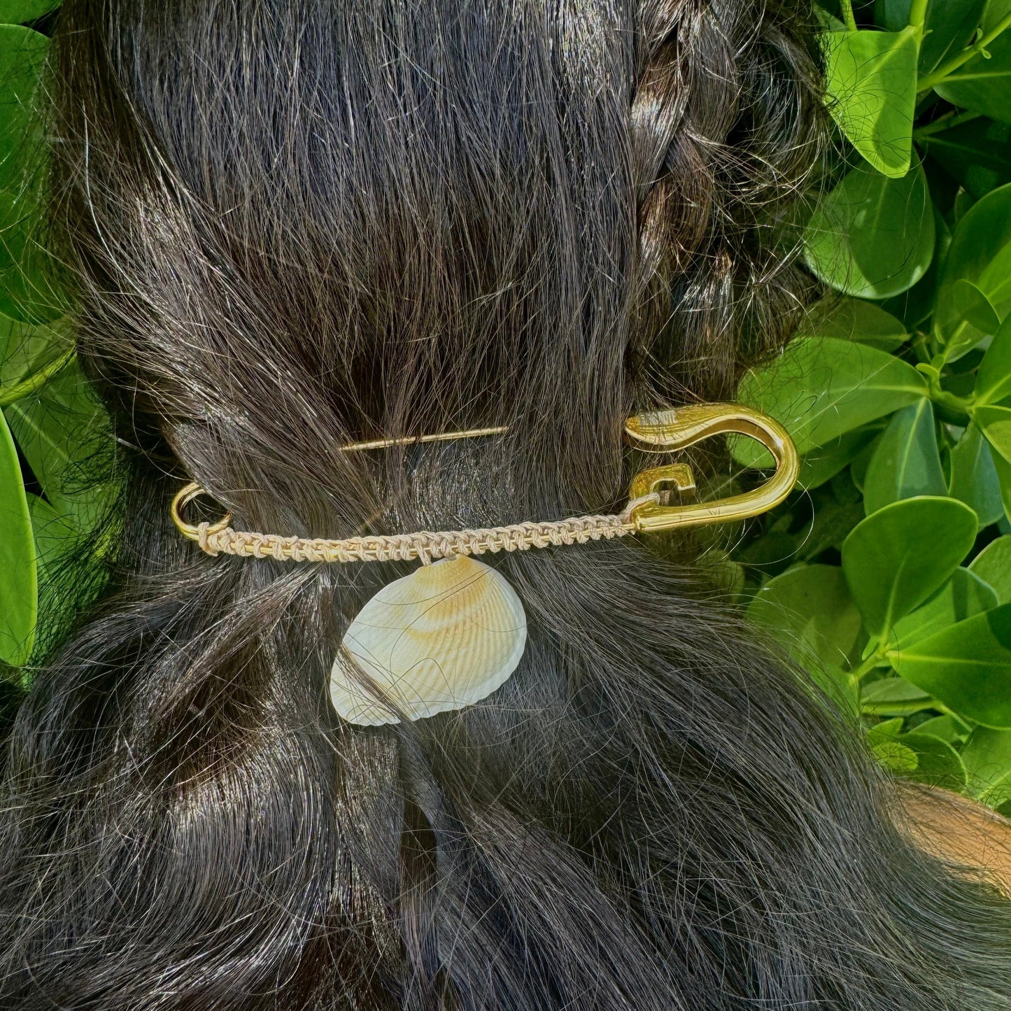 A close-up of a person's dark hair styled with a braided section and a gold-tone safety pin hair accessory holding a small white clam shell, set against green foliage.