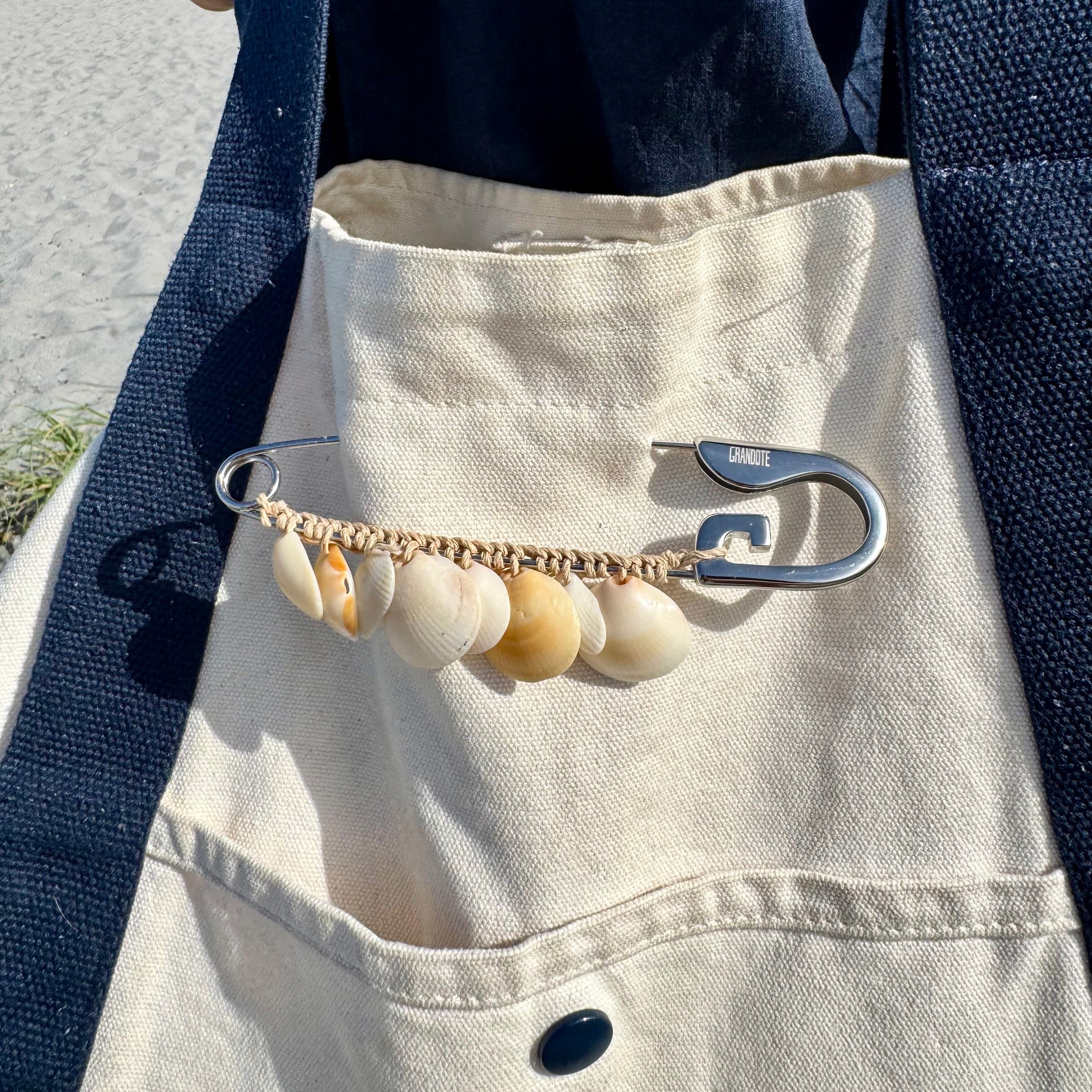 A close-up of a beige canvas tote bag with a front pocket, featuring a decorative chain accessory with shell charms and silver safety-pin style clips, worn across the pocket. The bag is shown outdoors on a beach with sand and ocean visible in the background.