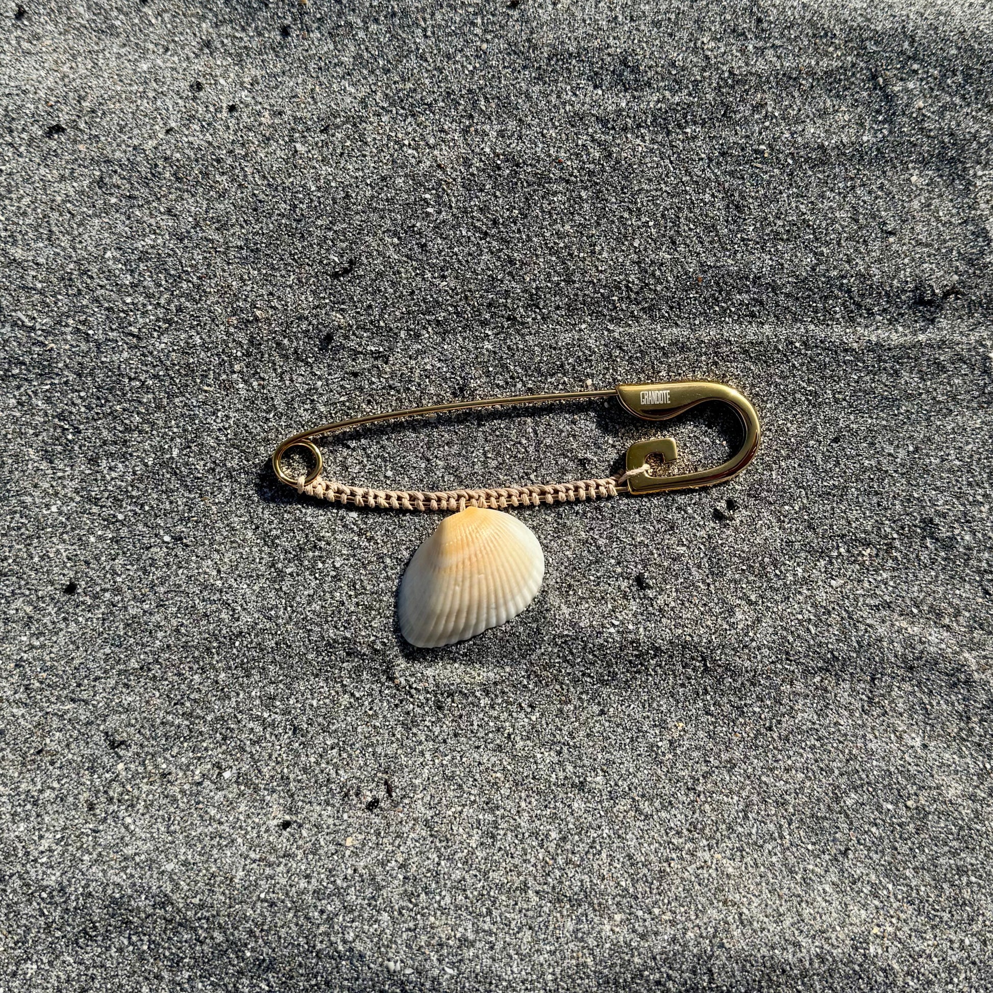 A small safety pin with a white seashell attached, lying on gray sand.