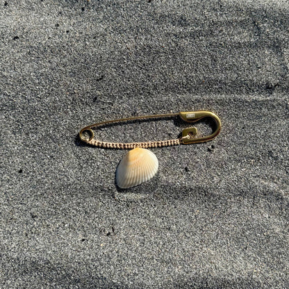 A small safety pin with a white seashell attached, lying on gray sand.