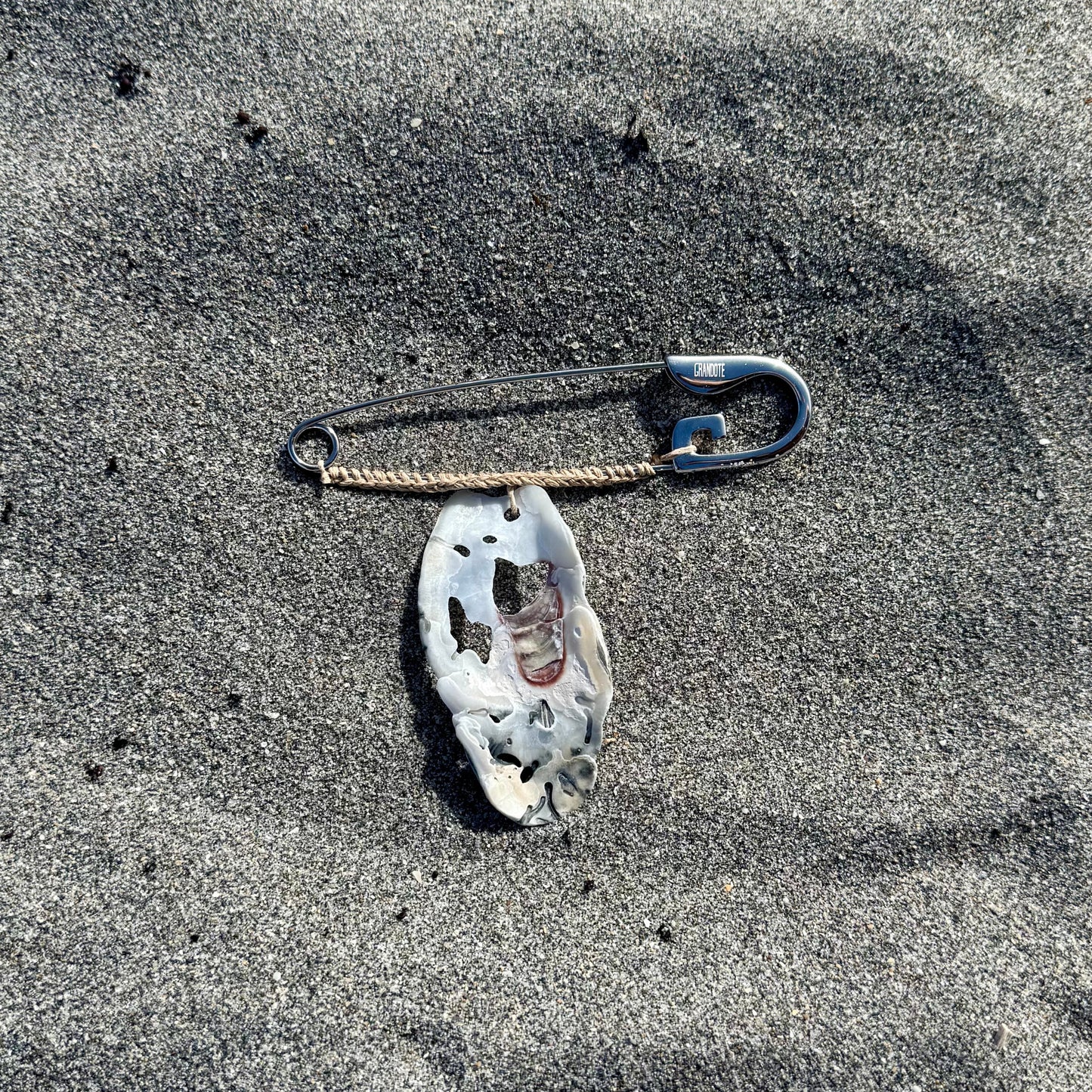 A large safety pin with a white, patterned oyster shell attached, lying on sand.