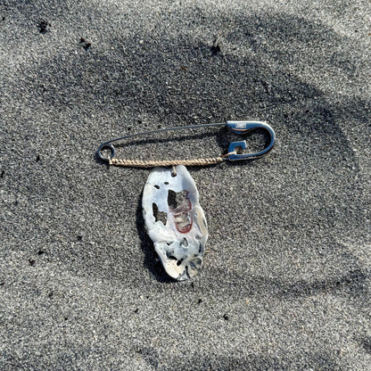 A large safety pin with a white, patterned oyster shell attached, lying on sand.