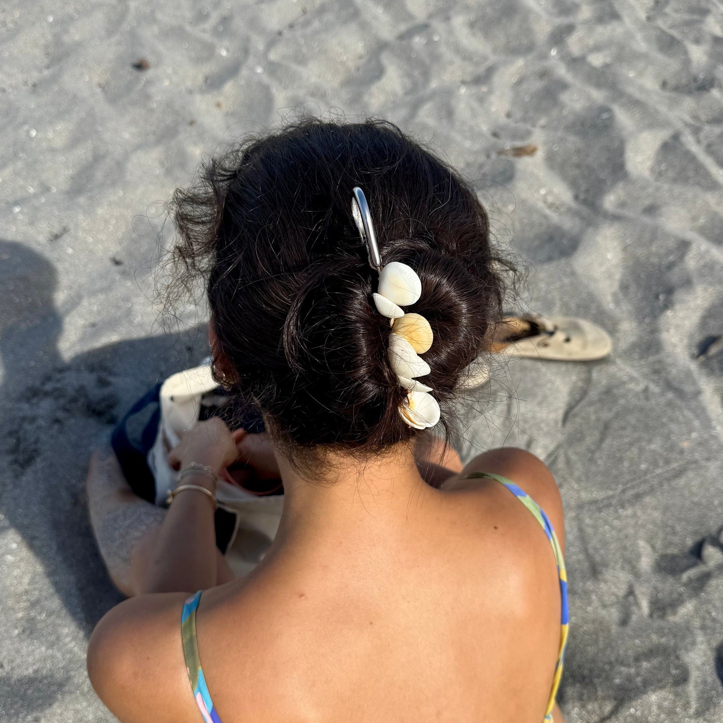A woman sitting on a sandy beach viewed from behind, wearing a colorful string bikini and a silver hair clip adorned with white seashells in her hair.