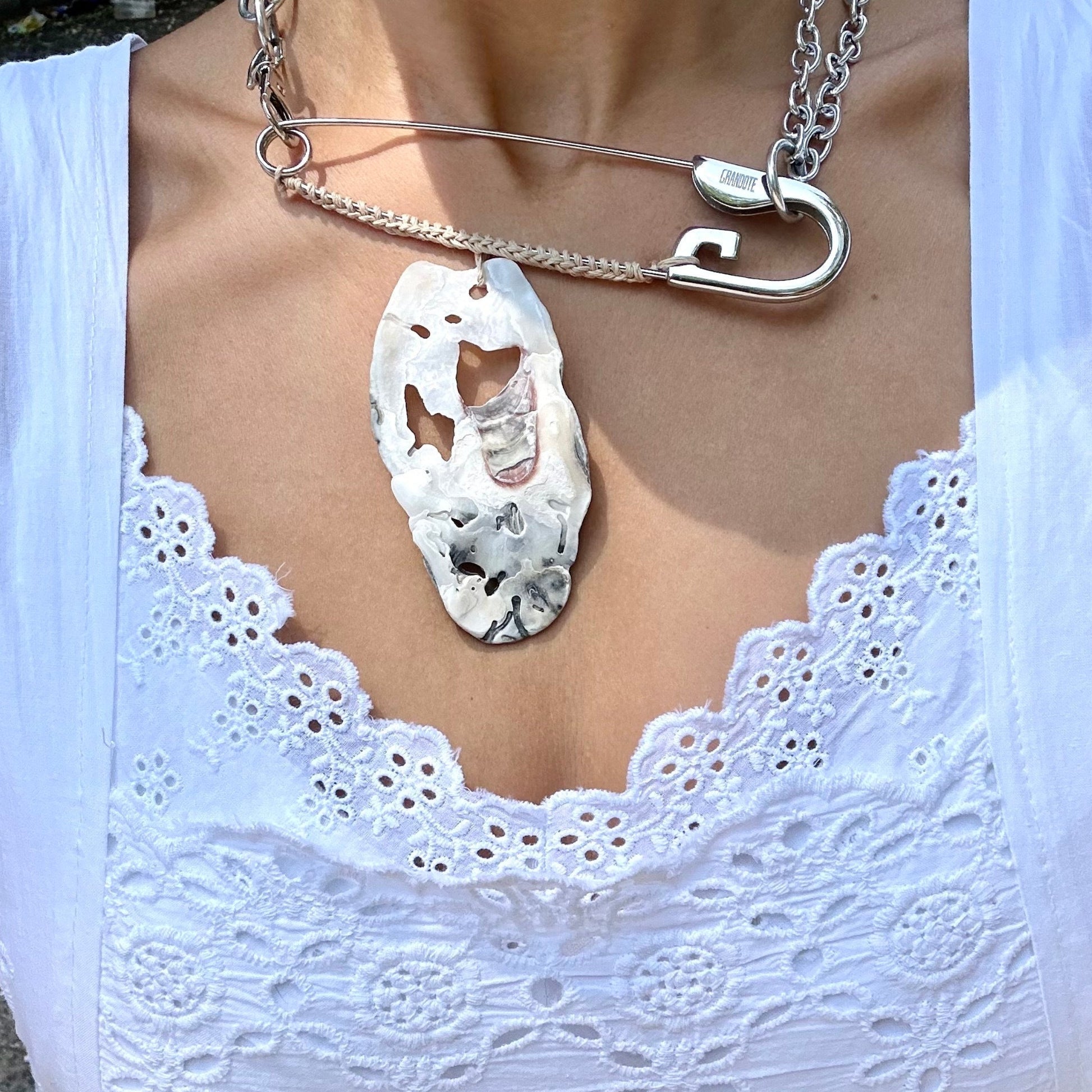 A close-up of a person wearing a white eyelet lace top and a silver necklace with a large, irregular white oyster shell pendant.