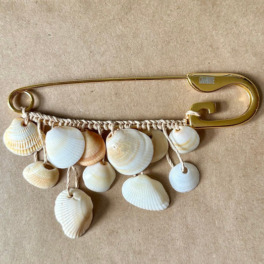 A gold safety pin brooch decorated with various small seashells and shell charms, displayed on a beige textured card.