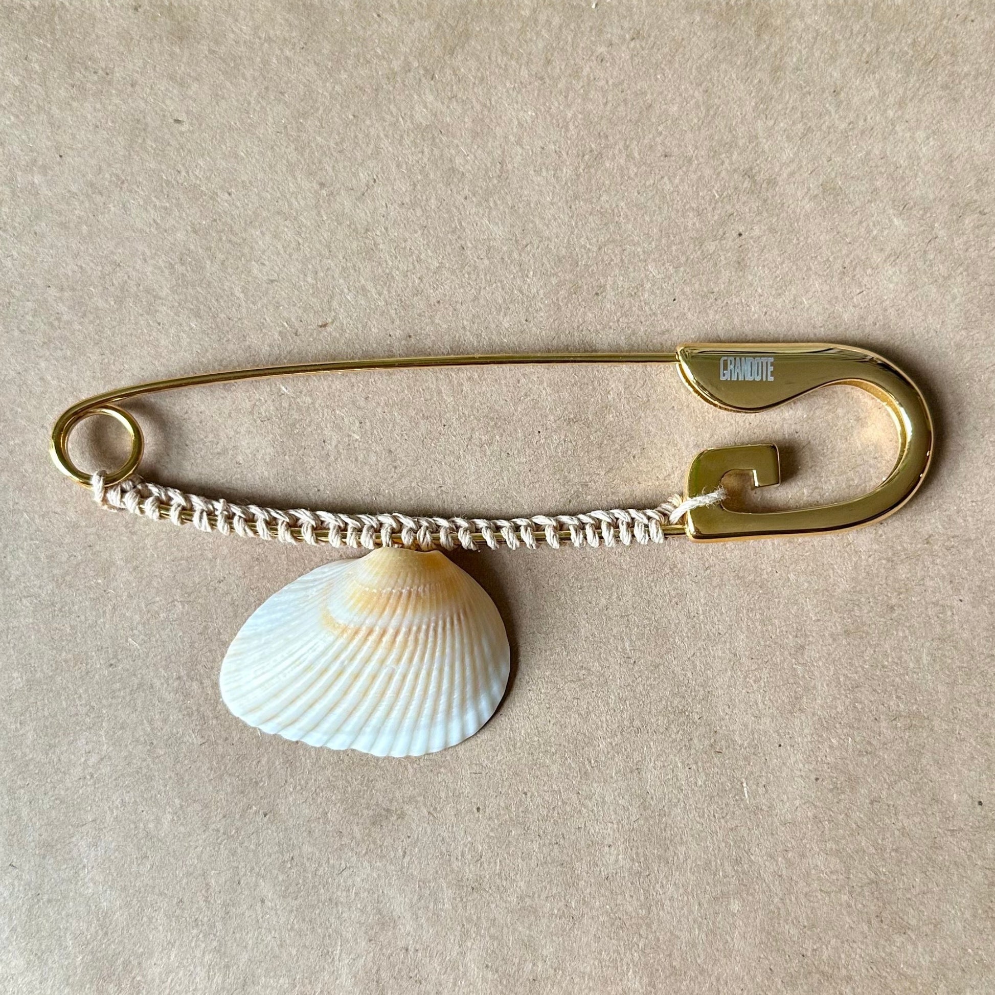 A gold-toned oversized safety pin accessory with a white clam shell charm, displayed on a beige textured surface.