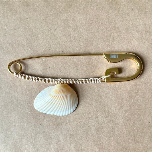 A gold-toned oversized safety pin accessory with a white clam shell charm, displayed on a beige textured surface.