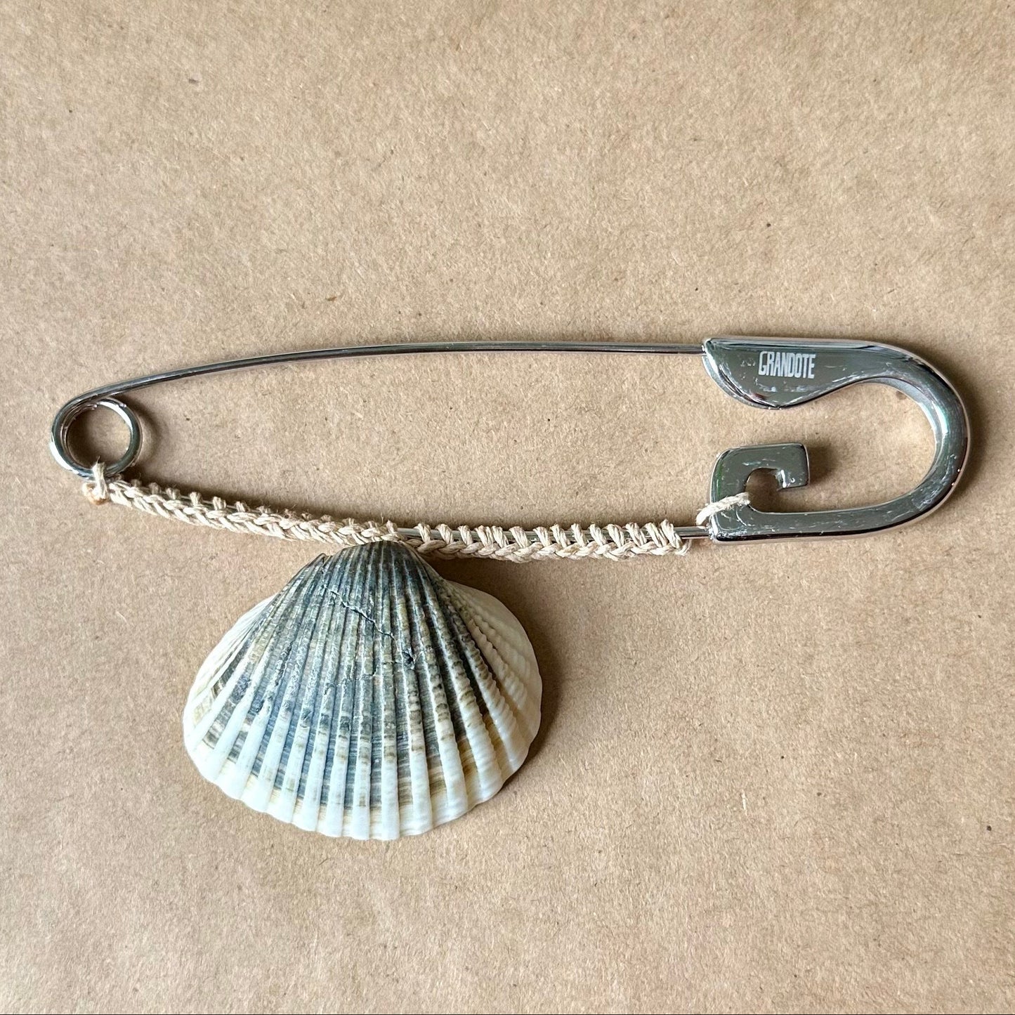 A silver safety pin with a grey seashell charm, displayed on a brown kraft paper sheet.
