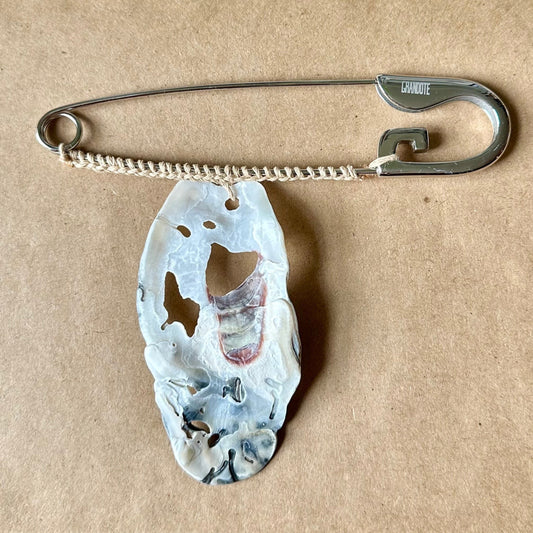 A silver safety pin with a carved, irregularly shaped white and gray oyster shell pendant, displayed on a brown cardboard surface.