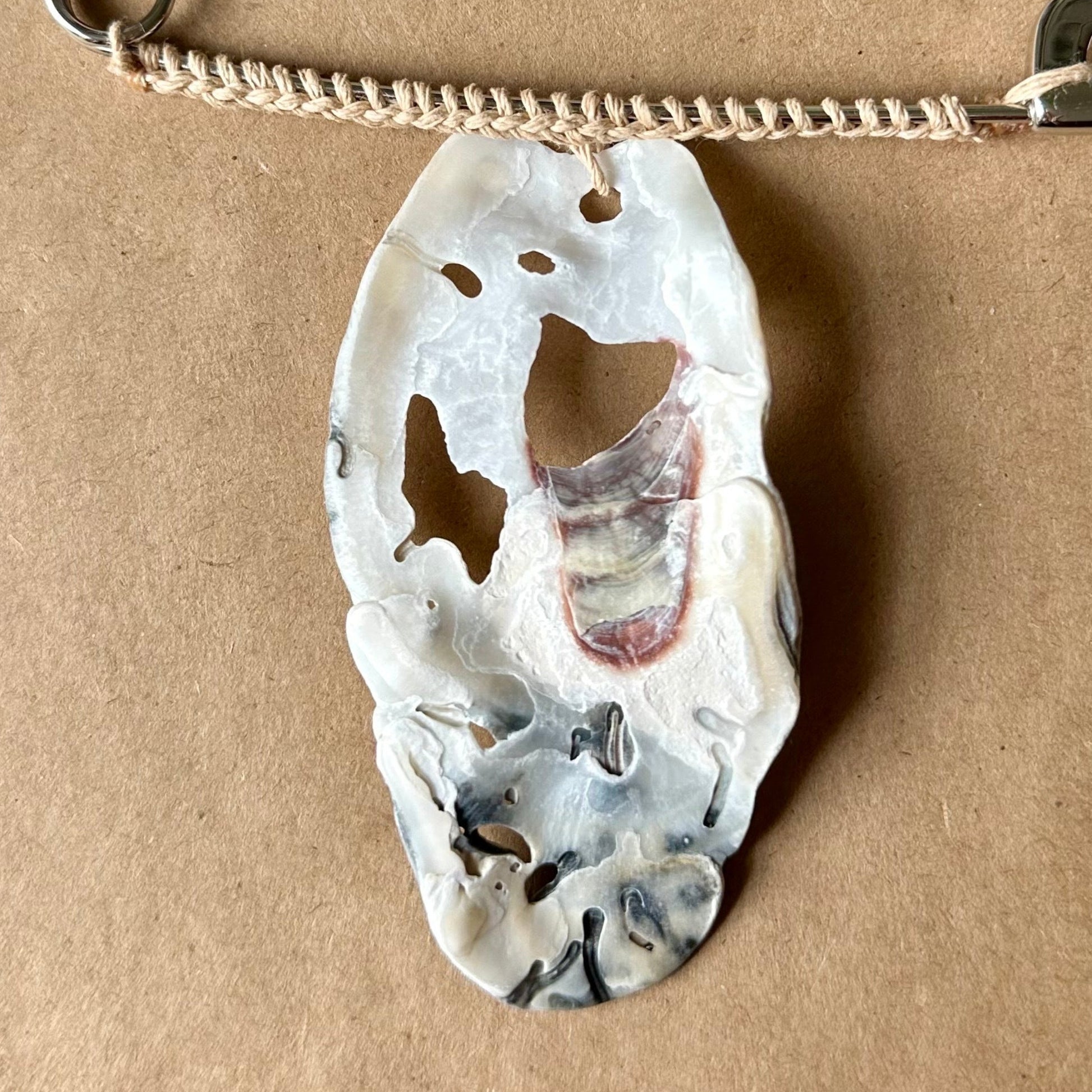 A silver safety pin necklace with a carved white, black, and red oyster shell pendant, displayed on a brown cardboard surface.