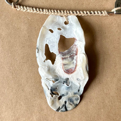 A silver safety pin necklace with a carved white, black, and red oyster shell pendant, displayed on a brown cardboard surface.