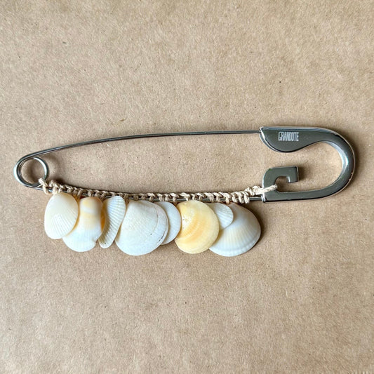 A silver safety pin styled as a brooch, decorated with a small chain of seashells in white, beige, and light yellow, resting on a brown textured paper surface.